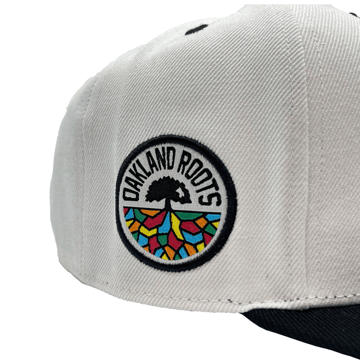 Close-up of the Oakland Roots SC Arched Text Snapback, a white hat featuring a colorful round patch on the side. The patch displays a tree with black roots and rainbow-colored geometric shapes below. The text "OAKLAND ROOTS" encircles the tree symbol in black capital letters, reminiscent of Oaklandish style. The bill of the cap is dark.