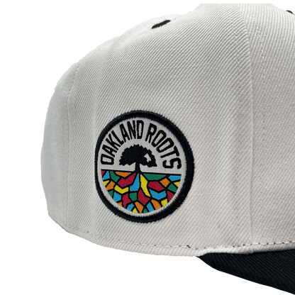 Close-up of the Oakland Roots SC Arched Text Snapback, a white hat featuring a colorful round patch on the side. The patch displays a tree with black roots and rainbow-colored geometric shapes below. The text "OAKLAND ROOTS" encircles the tree symbol in black capital letters, reminiscent of Oaklandish style. The bill of the cap is dark.