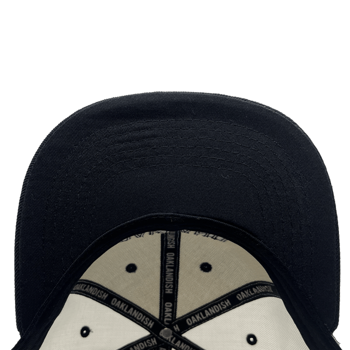 An underside view of the Oakland Roots SC Arched Text Snapback. The interior is lined with cream-colored fabric and accented by black stitching, along with a black band that repeatedly reads "OAKLANDISH." The cap's brim is prominently visible, displaying detailed stitching. The background is plain and white.