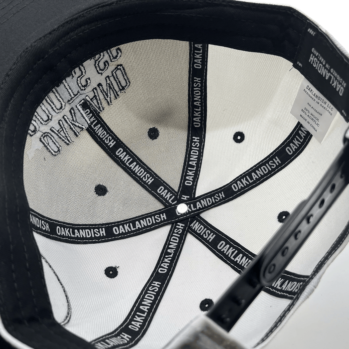 The image showcases the interior of an Oakland Roots SC Arched Text Snapback hat. The inside features white fabric sections printed with "OAKLANDISH" in black, and you can see black ventilation holes and stitching. There is an adjustable strap at the back, along with a white label bearing care instructions.
