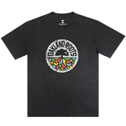 The Oakland Roots SC Classic Heavy Stone Wash Tee is a black, short-sleeved T-shirt that features a central circular logo. The logo showcases an abstract tree design with vibrant, geometric leaves and the text "Oakland Roots" arched above it, symbolizing the sports club's dedication to social good. The Oaklandish brand logo is printed near the collar for an added touch of authenticity.