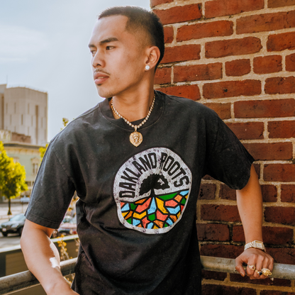 A young man with short hair stands on a balcony by a brick wall, wearing the Oakland Roots SC Classic Heavy Stone Wash Tee with a colorful logo. He accessorizes with gold jewelry, looking off to the side; city and trees are softly blurred behind him.