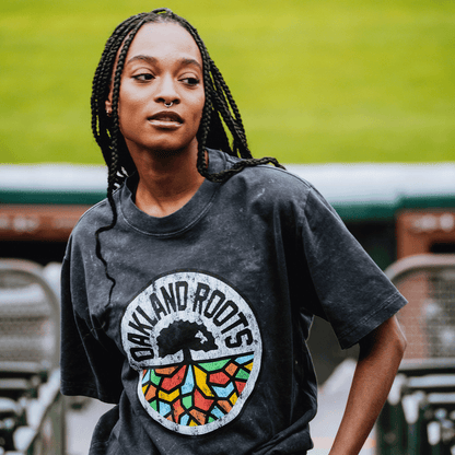 A woman with long braids and a nose ring wears the Oakland Roots SC Classic Heavy Stone Wash Tee by Oakland Roots SC, featuring a tree and colorful roots. She stands outside near stadium seats, with a blurred green field behind her.