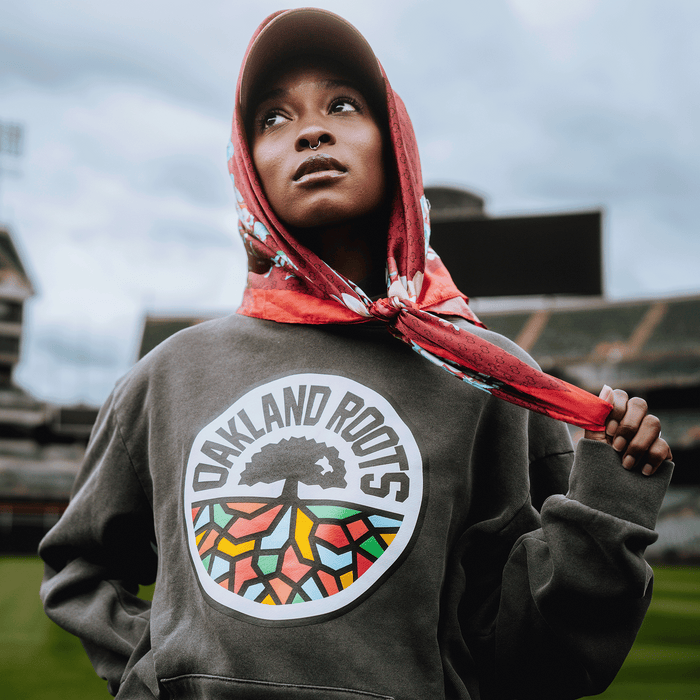 A woman in a red scarf and gray Oakland Roots SC Heavy Hoodie stands in a stadium, gazing upward with confidence. The hoodie, by Oakland Roots SC, displays a tree logo with colorful geometric roots.