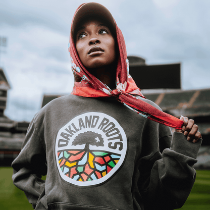 A woman in a red scarf and gray Oakland Roots SC Heavy Hoodie stands in a stadium, gazing upward with confidence. The hoodie, by Oakland Roots SC, displays a tree logo with colorful geometric roots.