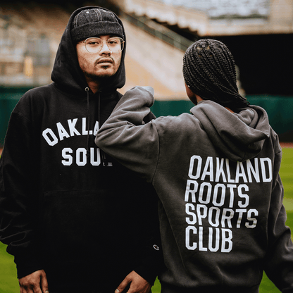 Two people stand on a sports field in hoodies. One, facing forward in glasses, wears a black Oaklandish Pullover Hoodie; the other, with braided hair and back turned, is in an Oakland Roots SC Heavy Hoodie. Stadium seating appears blurred behind them.
