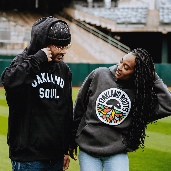 Two people smile at each other on a baseball field. One wears an Oaklandish “OAKLAND SOUL” hoodie, while the other sports an Oakland Roots SC Heavy Hoodie by Oakland Roots SC—each proudly repping their favorite local club.