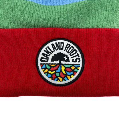 A close-up of the Oakland Roots SC Mosaic Stripe Beanie shows a textured fabric with green on top and red on the bottom. In the center, a circular patch features a stylized tree with roots spreading out, colored in vibrant, mosaic-like patterns. The words "OAKLAND ROOTS Sports Club" are displayed in a semicircle above the tree.