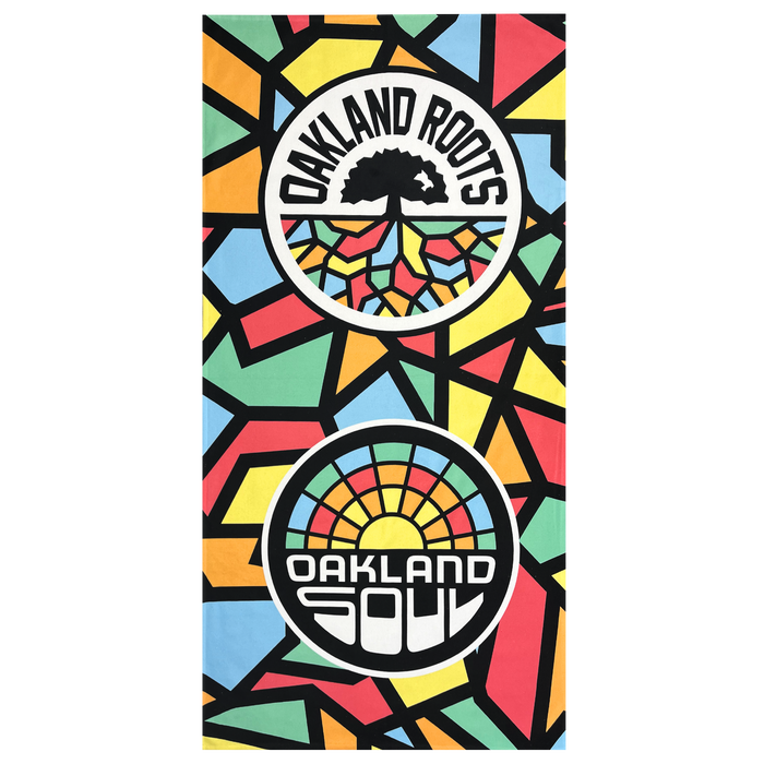 The Oakland Roots SC & Soul Towel by Oakland Roots SC is a brightly colored, premium dye-sublimated beach towel featuring a geometric mosaic pattern. It showcases two circular logos: one with a black tree and the text "OAKLAND ROOTS," and the other with a sunrise and the text "OAKLAND SOUL." The background displays vibrant shapes in red, yellow, green, blue, and black. Proceeds from this product support social good initiatives by the Oakland Roots Sports Club.
