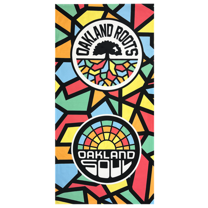 The Oakland Roots SC & Soul Towel by Oakland Roots SC is a brightly colored, premium dye-sublimated beach towel featuring a geometric mosaic pattern. It showcases two circular logos: one with a black tree and the text "OAKLAND ROOTS," and the other with a sunrise and the text "OAKLAND SOUL." The background displays vibrant shapes in red, yellow, green, blue, and black. Proceeds from this product support social good initiatives by the Oakland Roots Sports Club.