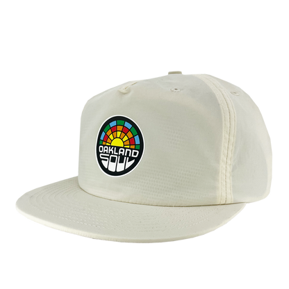 The Oakland Soul Surf Cap is a cream-colored baseball cap with a flat brim, featuring a colorful circular logo on the front. The logo displays a multicolored rainbow-like arc above the text "Oakland Soul," symbolizing the dynamic women's soccer team in the USL W-League, written in a bold, white, modern font against a black background.