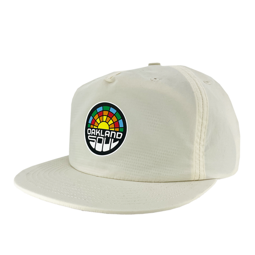 The Oakland Soul Surf Cap is a cream-colored baseball cap with a flat brim, featuring a colorful circular logo on the front. The logo displays a multicolored rainbow-like arc above the text "Oakland Soul," symbolizing the dynamic women's soccer team in the USL W-League, written in a bold, white, modern font against a black background.