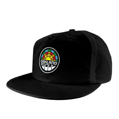 The Oakland Soul Surf Cap is a black snapback hat boasting a vibrant geometric design reminiscent of a sunrise or sunset on the front panel. Below this design, the text "OAKLAND SOUL" is prominently displayed in honor of the women's soccer team from the USL W-League. The hat features a slightly curved bill and noticeable stitching details.