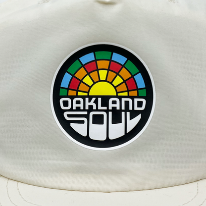 The Oakland Soul Surf Cap is a stylish white hat featuring a circular logo with a yellow sun at its center, surrounded by blue, green, orange, and red hues that create an eye-catching stained glass effect. Beneath this vibrant design, "OAKLAND SOUL" is boldly printed in modern white letters against a black background. Show your support for the Oakland Soul USL W-League women's soccer team in style with this fashionable accessory.