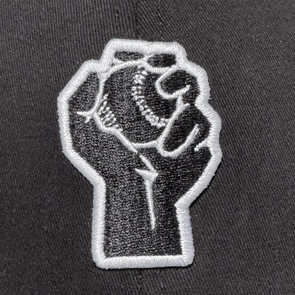 The Oakland Stand Up Hat by Oakland Ballers features a black and white embroidered patch of a raised fist holding a baseball, symbolizing solidarity and the spirit of the city’s newest Pioneer League team.