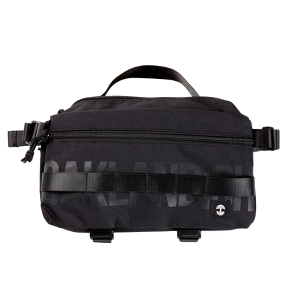 A black nylon hip bag with a black Oaklandish wordmark, front zipper, top handle, waist belt, & small white Oaklandish tree logo.
