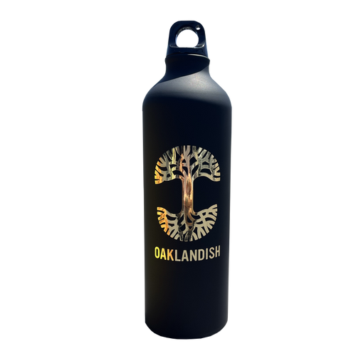 The Oaklandish Logo Aluminum Water Bottle by Oaklandish is crafted from single-wall aluminum and features a striking black exterior. The front showcases the iconic Oaklandish tree logo nestled within a circular "C" shape, along with "OAKLANDISH" boldly displayed beneath in uppercase white letters. It comes with a convenient looped handle cap for easy carrying, set against a plain white background.