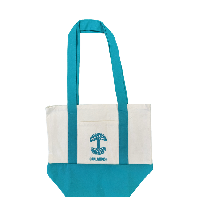 The Oaklandish Mini Grocery Tote by Oaklandish features teal blue handles and bottom with a white center. An embroidered turquoise tree logo and the word “OAKLANDISH” decorate the front pocket. The tote is shown on a plain white background.