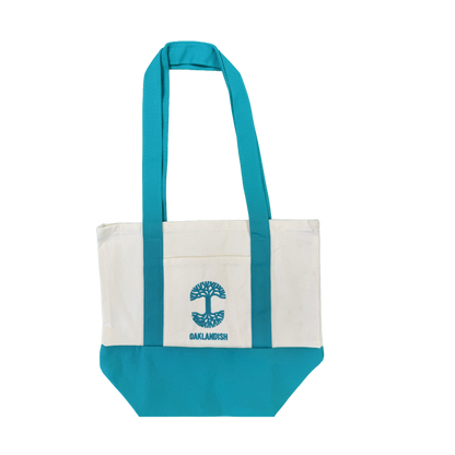 The Oaklandish Mini Grocery Tote by Oaklandish features teal blue handles and bottom with a white center. An embroidered turquoise tree logo and the word “OAKLANDISH” decorate the front pocket. The tote is shown on a plain white background.