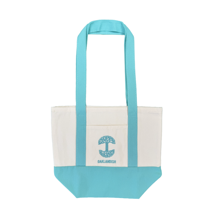 Introducing the Oaklandish Mini Grocery Tote by Oaklandish—a canvas tote bag featuring aqua blue handles and bottom, a white body, and a convenient front pocket. The pocket is adorned with an embroidered aqua blue tree graphic and “OAKLANDISH” written below. Ideal for use as a mini grocery tote or carrying your everyday essentials. Shown against a plain white background.