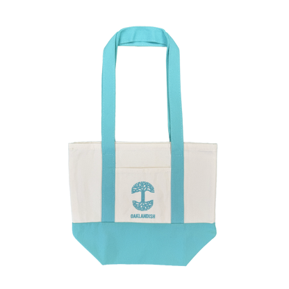 Introducing the Oaklandish Mini Grocery Tote by Oaklandish—a canvas tote bag featuring aqua blue handles and bottom, a white body, and a convenient front pocket. The pocket is adorned with an embroidered aqua blue tree graphic and “OAKLANDISH” written below. Ideal for use as a mini grocery tote or carrying your everyday essentials. Shown against a plain white background.