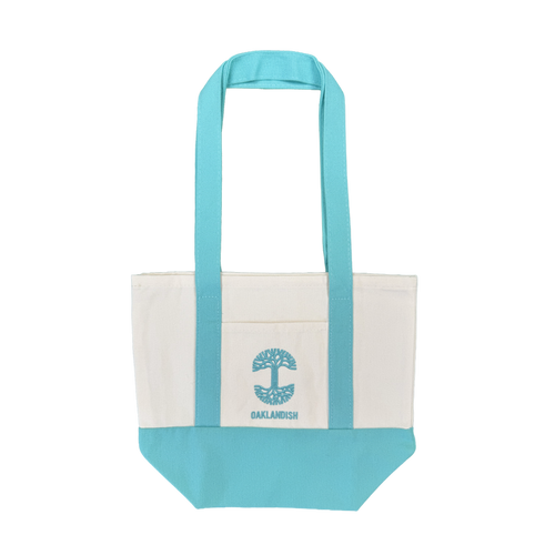 Introducing the Oaklandish Mini Grocery Tote by Oaklandish—a canvas tote bag featuring aqua blue handles and bottom, a white body, and a convenient front pocket. The pocket is adorned with an embroidered aqua blue tree graphic and “OAKLANDISH” written below. Ideal for use as a mini grocery tote or carrying your everyday essentials. Shown against a plain white background.