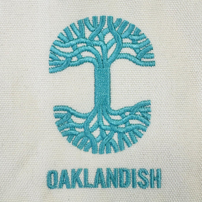 Featuring an eye-catching embroidered logo, the Oaklandish Mini Grocery Tote showcases a teal abstract oak tree forming a circular design above the all-caps “OAKLANDISH” brand name, stitched on textured cream fabric for standout style.