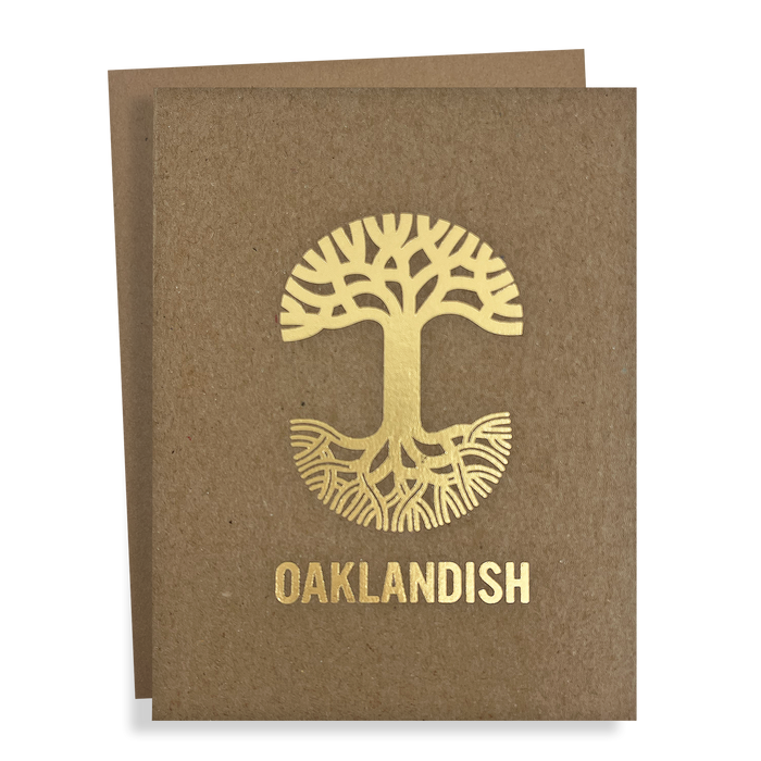 The "Oaklandish New Logo Card" by Never Elsewhere is a greeting card made from recycled brown kraft paper. It features a golden, stylized tree with roots extending in a circular design, and prominently displays the text "OAKLANDISH" below the tree. Another card slightly peeks out from behind, suggesting it's part of a set.