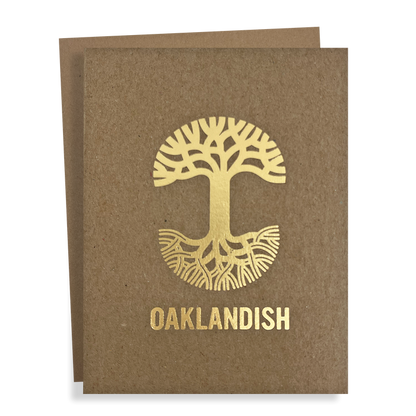 The "Oaklandish New Logo Card" by Never Elsewhere is a greeting card made from recycled brown kraft paper. It features a golden, stylized tree with roots extending in a circular design, and prominently displays the text "OAKLANDISH" below the tree. Another card slightly peeks out from behind, suggesting it's part of a set.