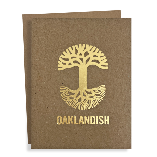 The "Oaklandish New Logo Card" by Never Elsewhere is a greeting card made from recycled brown kraft paper. It features a golden, stylized tree with roots extending in a circular design, and prominently displays the text "OAKLANDISH" below the tree. Another card slightly peeks out from behind, suggesting it's part of a set.