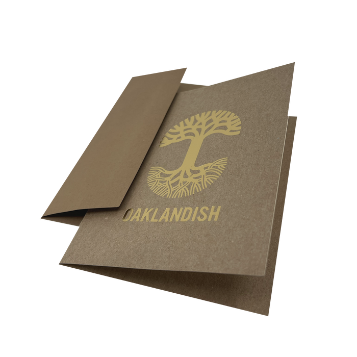 An "Oaklandish New Logo Card" by Never Elsewhere, crafted from recycled brown kraft paper, lies partially open on a white background. The card displays a stylized gold tree with exposed roots and the word "OAKLANDISH" printed beneath it using letterpress print. One flap is partially folded inward, revealing the inside of the card.