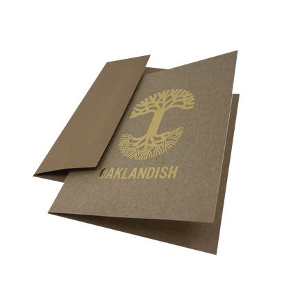 An "Oaklandish New Logo Card" by Never Elsewhere, crafted from recycled brown kraft paper, lies partially open on a white background. The card displays a stylized gold tree with exposed roots and the word "OAKLANDISH" printed beneath it using letterpress print. One flap is partially folded inward, revealing the inside of the card.