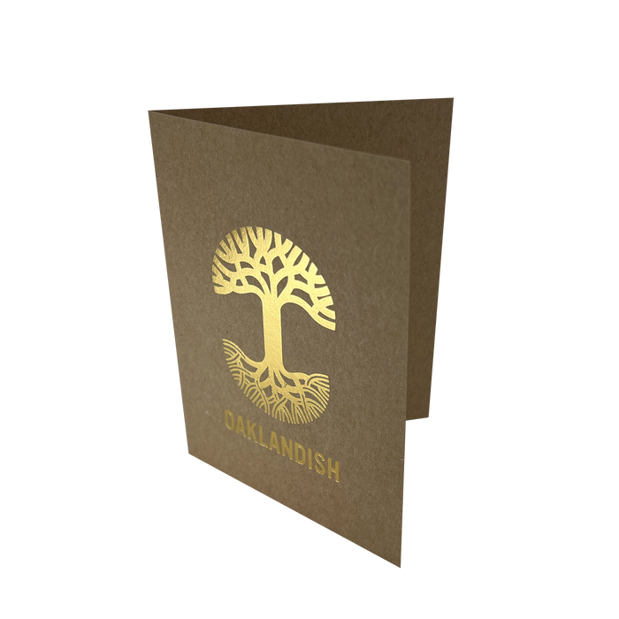 The Oaklandish New Logo Card by Never Elsewhere is a minimalist greeting card made of recycled brown kraft paper, shown partially open. The front showcases a gold tree with intricate roots and branches, printed using letterpress. Below the tree, "OAKLANDISH" is displayed in gold capital letters. The card rests on a white background.