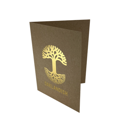 The Oaklandish New Logo Card by Never Elsewhere is a minimalist greeting card made of recycled brown kraft paper, shown partially open. The front showcases a gold tree with intricate roots and branches, printed using letterpress. Below the tree, "OAKLANDISH" is displayed in gold capital letters. The card rests on a white background.