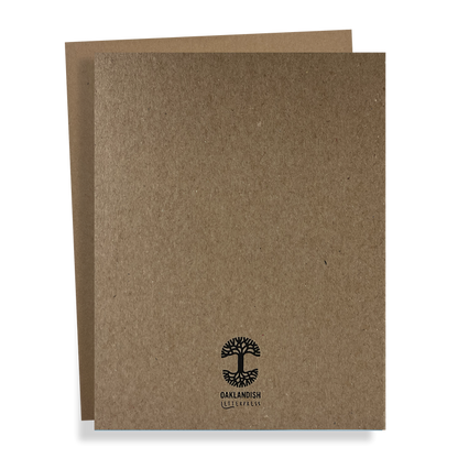 Two brown cardboard folders are stacked, with the one in front slightly overlapping the one at the back. The front folder, known as the Oaklandish New Logo Card by Never Elsewhere, is crafted from recycled brown kraft paper and features a black logo of a tree with roots along with the text "OAKLANDISH" underneath. The overall design is simple and minimalistic.