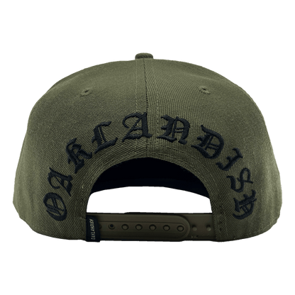 The Old English Aft Snapback by Oaklandish, showcased from the rear, features an adjustable strap and the word "OAKLANDISH" embroidered in a black Gothic-style font. The green cap includes a small black tag with white text near the strap and an Oaklandish tree logo. The fabric texture is also visible, highlighting its durable material.