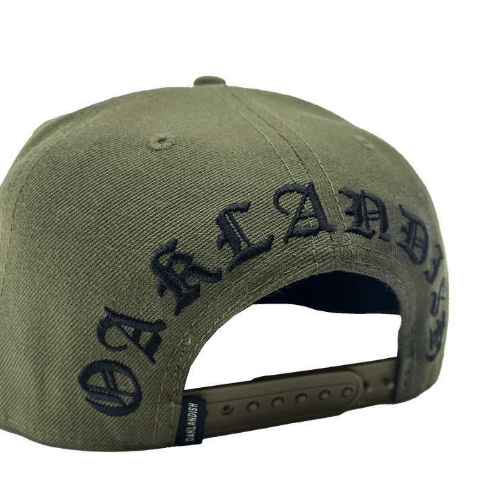 The Old English Aft Snapback by Oaklandish is an olive green hat featuring the word "OAKLANDISH" embroidered in black, gothic-style lettering across the back. This adjustable snapback has a dark green strap with multiple notches and a textured fabric for durability. The iconic Oaklandish tree logo adds a signature touch to this stylish cap.