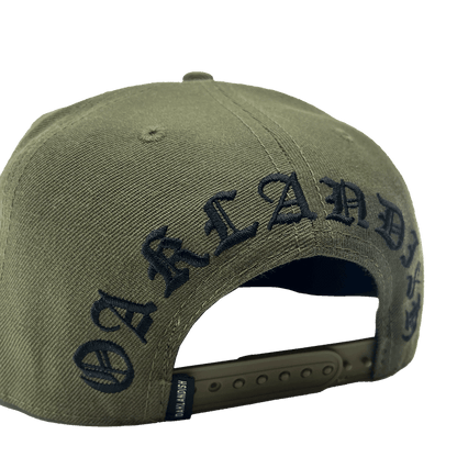 The Old English Aft Snapback by Oaklandish is an olive green hat featuring the word "OAKLANDISH" embroidered in black, gothic-style lettering across the back. This adjustable snapback has a dark green strap with multiple notches and a textured fabric for durability. The iconic Oaklandish tree logo adds a signature touch to this stylish cap.