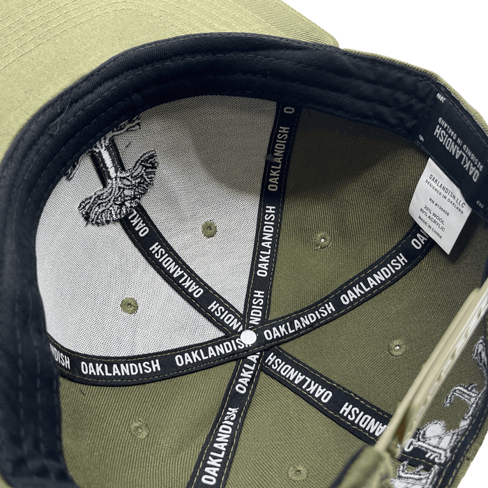 The image showcases the inside of an olive green "Old English Aft Snapback" hat by Oaklandish. The interior features black tape strips with the word "OAKLANDISH" repeatedly printed in white. The underside of the cap's brim displays a gray and white design. Additionally, a white label detailing care instructions is stitched into the lining, complementing its adjustable snapback feature.