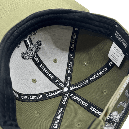 The image showcases the inside of an olive green "Old English Aft Snapback" hat by Oaklandish. The interior features black tape strips with the word "OAKLANDISH" repeatedly printed in white. The underside of the cap's brim displays a gray and white design. Additionally, a white label detailing care instructions is stitched into the lining, complementing its adjustable snapback feature.