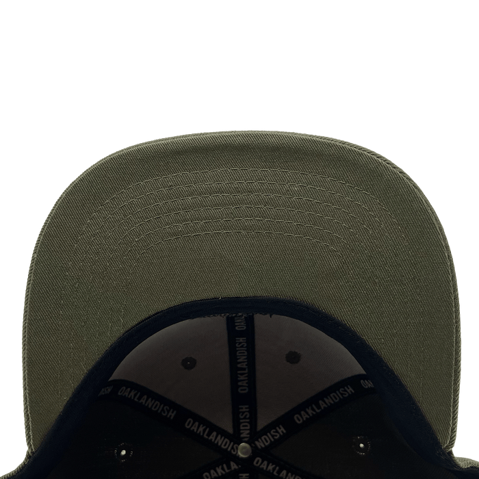 Close-up view of the underside of the Old English Aft Snapback's green brim. The stitching detail on the brim is clearly visible. The interior of this adjustable snapback cap by Oaklandish features a black lining with "OAKLANDISH" repeatedly printed in white on the inner bands. The background is plain white.