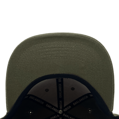Close-up view of the underside of the Old English Aft Snapback's green brim. The stitching detail on the brim is clearly visible. The interior of this adjustable snapback cap by Oaklandish features a black lining with "OAKLANDISH" repeatedly printed in white on the inner bands. The background is plain white.