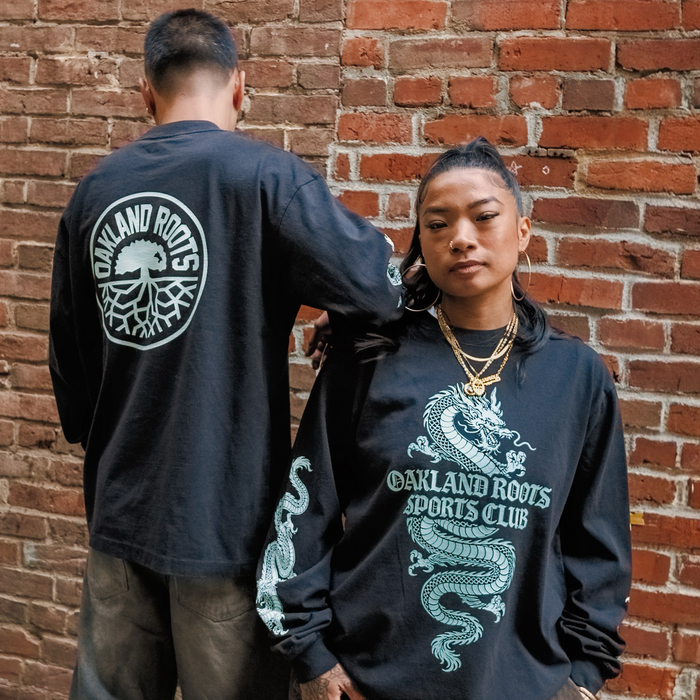 Two people pose confidently against a brick wall in matching black Oakland Roots SC ORSC Ryujin LS Tees, each featuring a white dragon design and the iconic roots logo on the back—showcasing bold style from Oakland Roots SC.