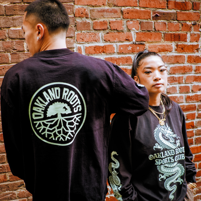 A man faces away, displaying an “Oakland Roots” logo on his shirt. A woman faces forward, wearing the Oakland Roots SC ORSC Ryujin LS Tee with a dragon sleeve design and club text on the front. Both stand by a brick wall.