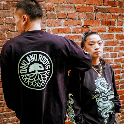 A man faces away, displaying an “Oakland Roots” logo on his shirt. A woman faces forward, wearing the Oakland Roots SC ORSC Ryujin LS Tee with a dragon sleeve design and club text on the front. Both stand by a brick wall.