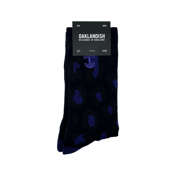A pair of Panther Socks featuring black crew length with blue polka dot and tree designs, packaged with a black label. The label reads "OAKLANDISH" with "DESIGNED IN OAKLAND" underneath. Additional text includes "TRD MRK" at the top and "EST. 2000" at the bottom. These Oaklandish logo socks are one size fits most.