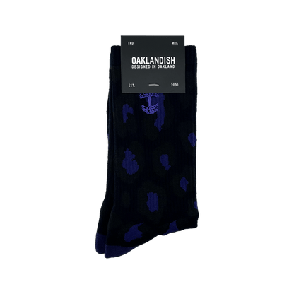 A pair of Panther Socks featuring black crew length with blue polka dot and tree designs, packaged with a black label. The label reads "OAKLANDISH" with "DESIGNED IN OAKLAND" underneath. Additional text includes "TRD MRK" at the top and "EST. 2000" at the bottom. These Oaklandish logo socks are one size fits most.