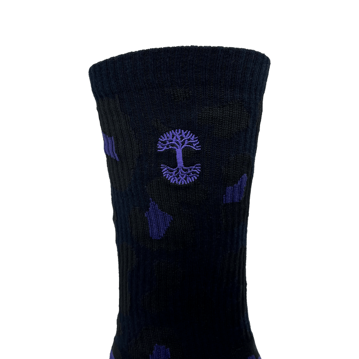 The Panther Sock by Oaklandish is displayed against a solid green background, showcasing its dark color with the Oaklandish logo prominently featured in the center. This one-size-fits-most crew sock has a ribbed texture, with the emblem outlined in purple within a circular design and slight purple accents scattered throughout.