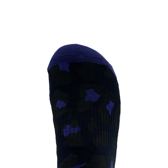 A close-up image of a sock-covered foot showcases Oaklandish's Panther Sock. The sock is primarily black with a dark blue toe cap and features a scattered pattern of blue spots. The plain white background emphasizes the design and colors of these one size fits most crew socks.