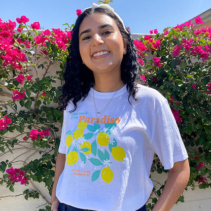 Smiling woman wearing the Paradiso T-Shirt with bright lemon and leaf design against vibrant pink flowers outdoors